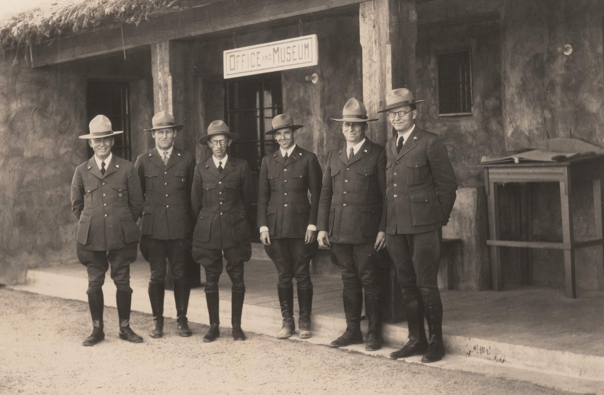 Six men stand in NPS uniforms with broad brim hats. Frank Pinkley wears a round superintendents badge and another ranger wears a shield-shaped badge.